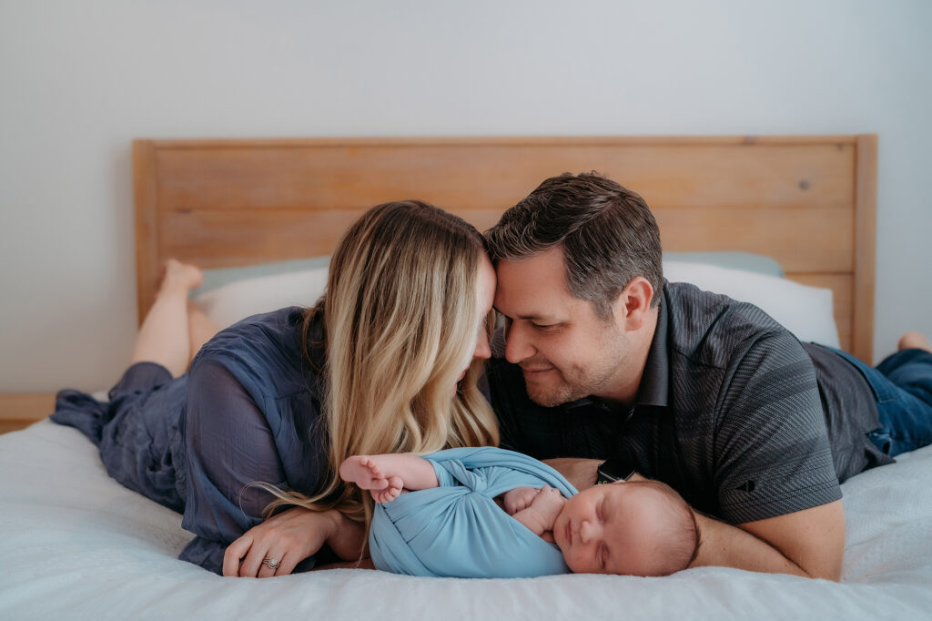 Denver Family Photographer captures mother and father laying in bed touching foreheads while watching baby