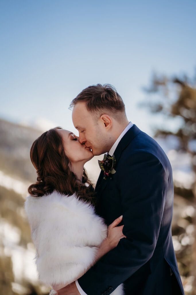 Groom holds his bride close as they embrace in the snow, the mountains and sun wrapping the moment in magic.