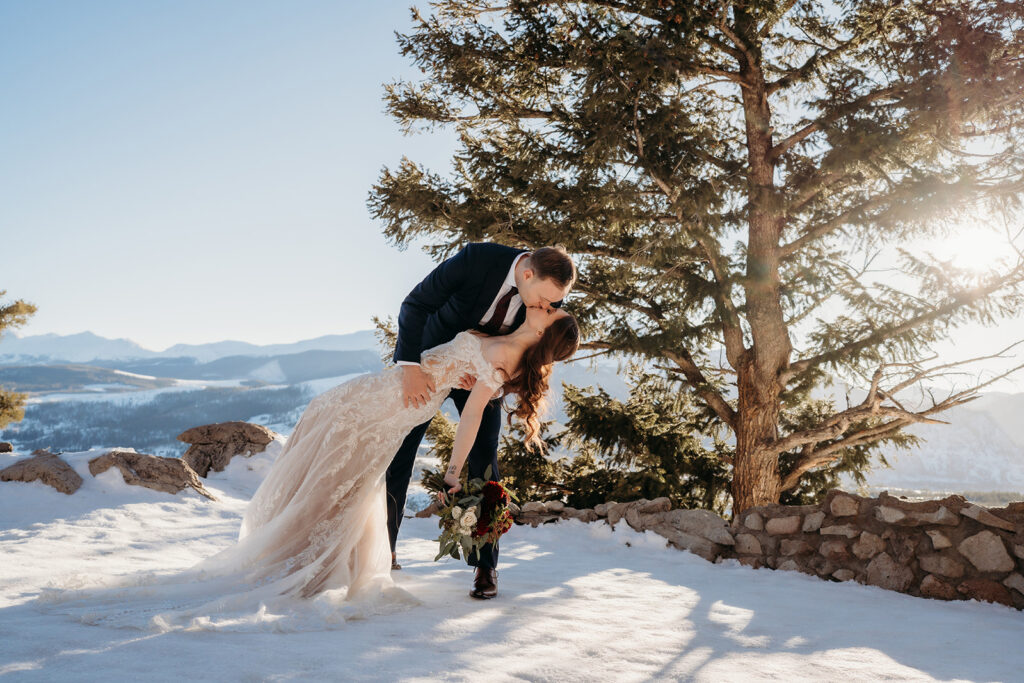 Groom dips and kisses his bride in the snow with mountain peaks behind them, capturing the best day of your life.