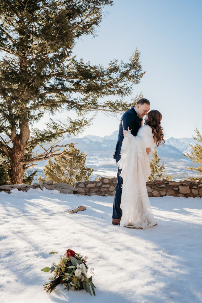 A couple kisses beneath a towering pine, their bouquet resting softly in the snow—Colorado elopement photographer.