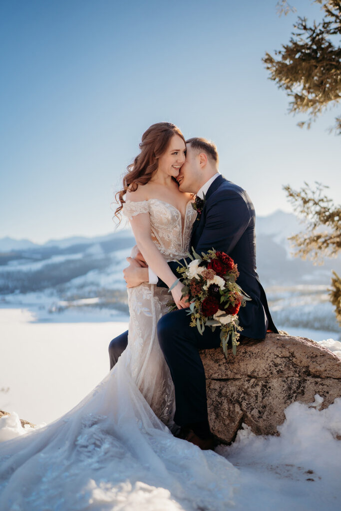 Bride sits on a snow-dusted boulder with her groom, laughing together in the sunlight with mountains behind them.