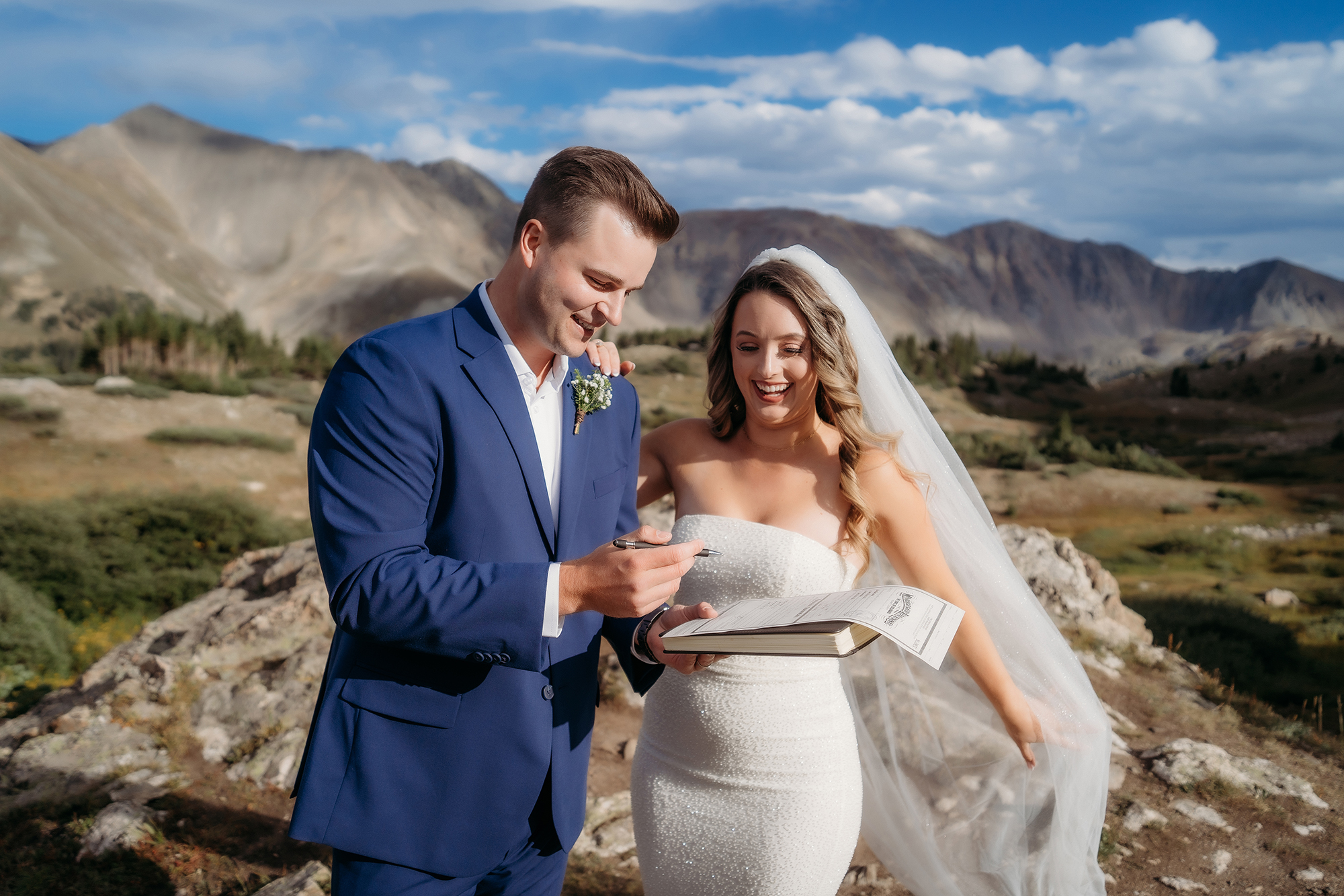 Groom signs the marriage license while the bride watches, both beaming under blue skies — Colorado Elopement Checklist.