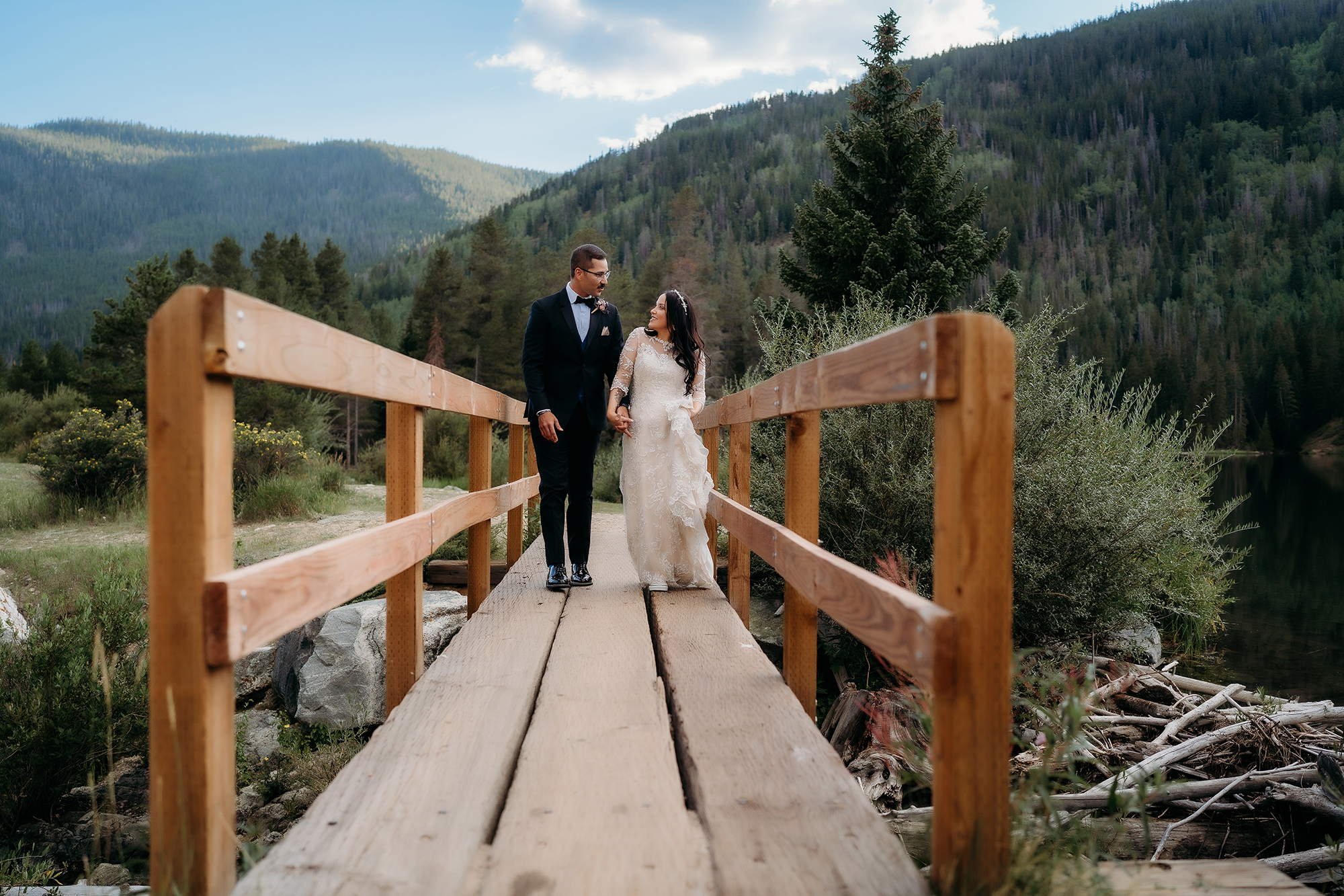 Bride and groom embrace on a small wooden bridge surrounded by alpine forest.