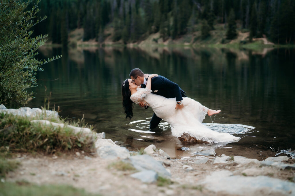 Newlyweds share a dip and kiss in a mountain lake, her lace dress trailing in the water.