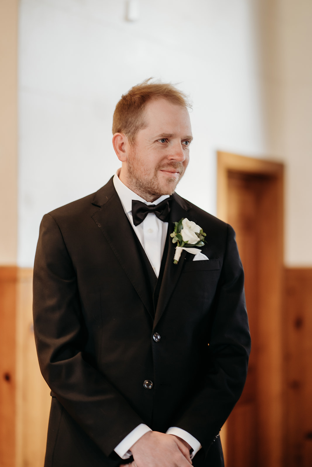 Groom smiles softly as he watches his bride approach the altar, framed by natural light in the chapel.