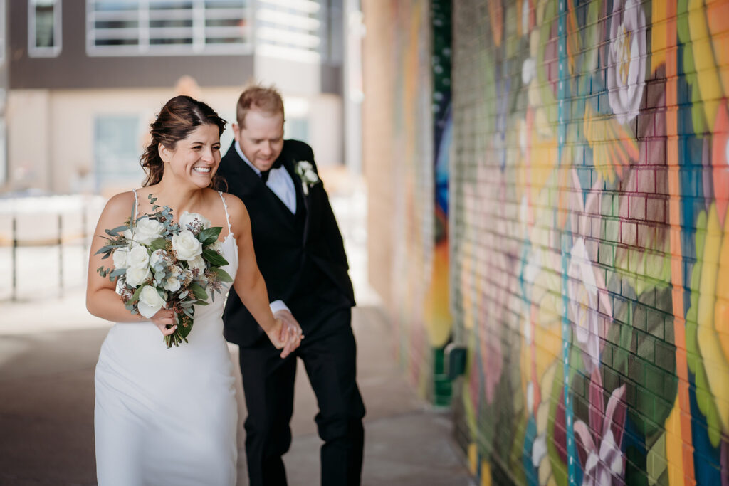 Newlyweds laugh and hold hands as they stroll through RiNo, celebrating their small wedding in Colorado with playful energy.