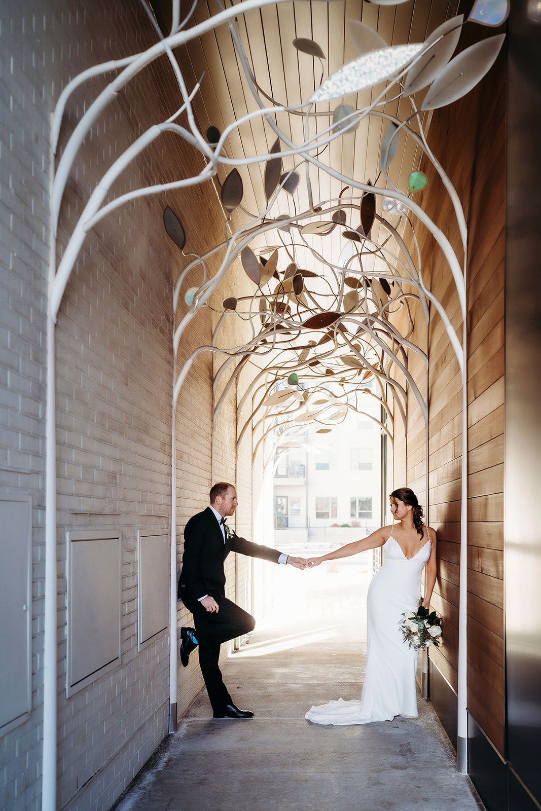 Bride and groom reach for each other across a modern urban archway, capturing a playful and romantic moment from their small wedding in Colorado.