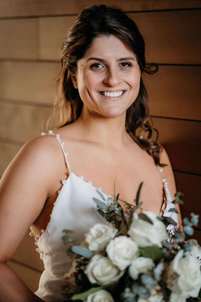 Bride smiles warmly, bouquet in hand, framed against wood paneling that highlights the elegance of her dress and flowers.