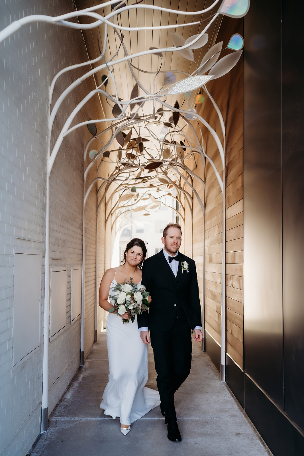 Newlyweds walk hand in hand beneath a sculptural metal archway, glowing in the late afternoon light after their small wedding in Colorado.