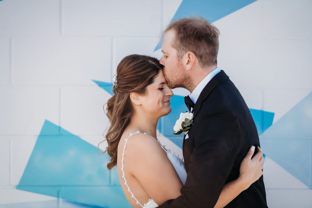 Groom kisses bride’s forehead as they embrace in front of a soft blue mural, wrapped in the quiet joy of the moment.
