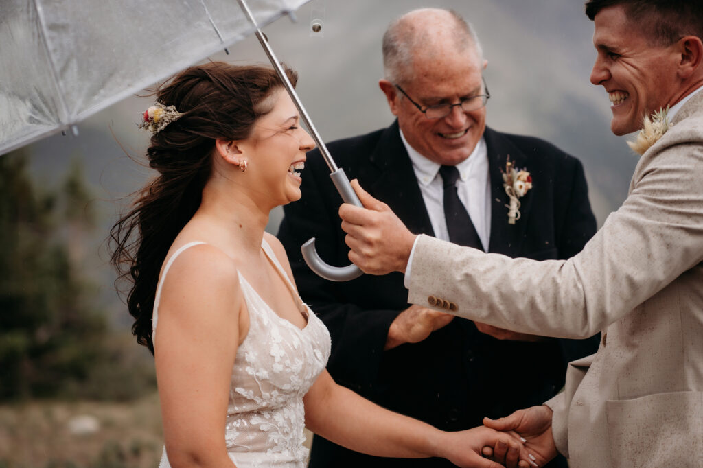 Bride and groom laugh under a clear umbrella during a rainy mountain elopement ceremony.