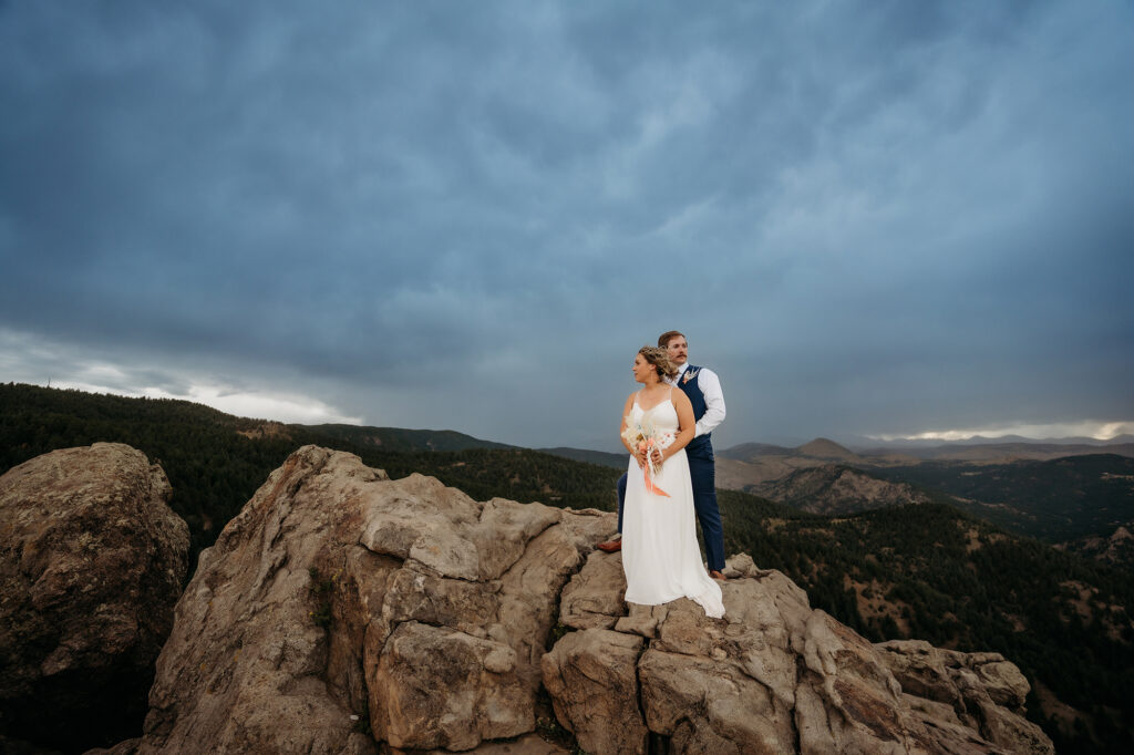 Couple stands confidently on rugged boulders overlooking a dramatic mountain landscape — Colorado Elopement Checklist.