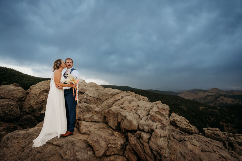 Bride kisses groom’s cheek as they stand on rocky cliffs, with the Colorado mountains behind them — Colorado Elopement Checklist.