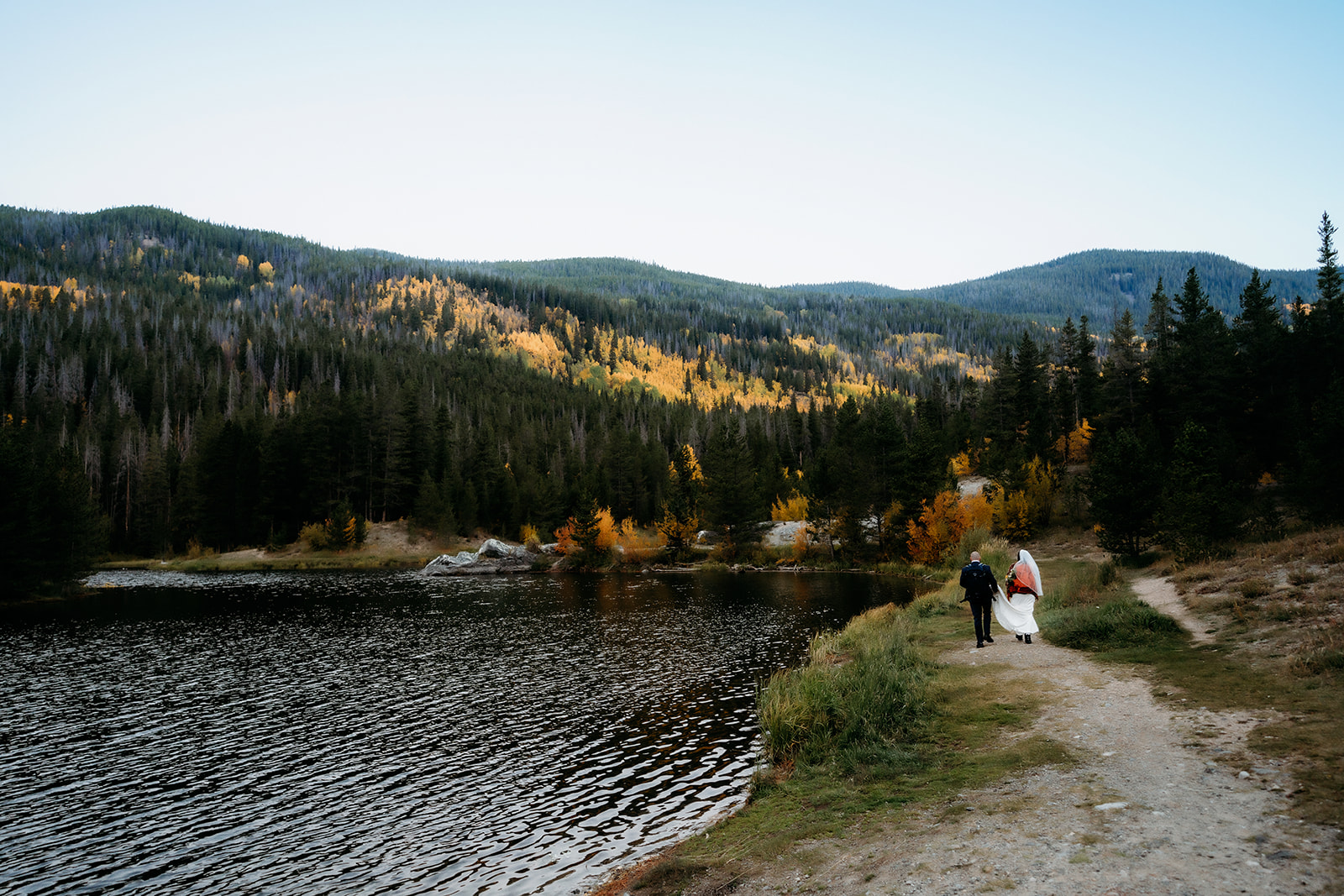 The bride and groom walk hand in hand along a peaceful trail beside a lake, surrounded by pine trees and glowing autumn foliage during their adventure elopement.