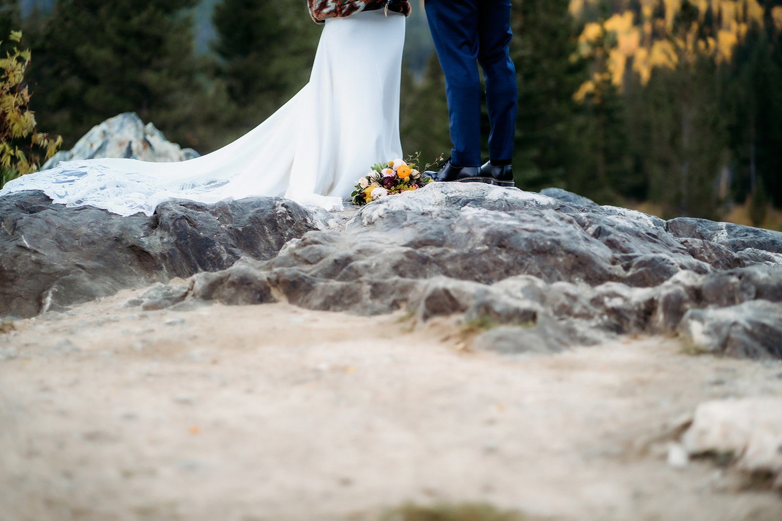 The train of the bride’s gown flows across rugged stone as their feet touch near a bouquet—capturing the texture and earthy beauty of a mountaintop elopement.