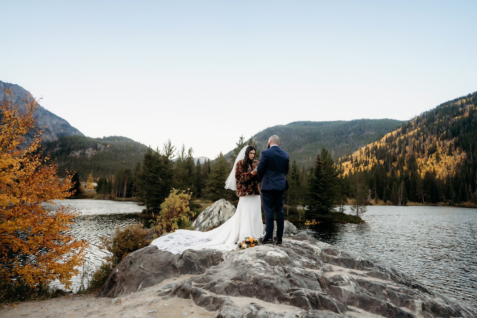 A bride and groom stand on a rocky outcrop beside a serene alpine lake, exchanging vows as golden aspens and rugged peaks surround them—an unforgettable adventure elopement moment.