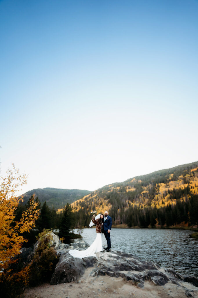 The bride raises her bouquet in the air with a big smile while standing beside her groom during their adventure elopement on a scenic Colorado mountain ledge.