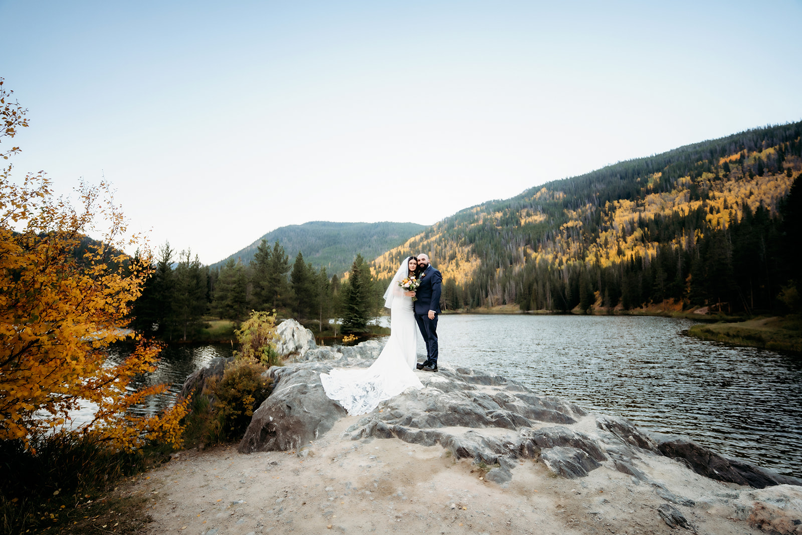 The couple embraces with joyful expressions, standing together on a rocky ledge above a calm lake framed by colorful fall trees and distant mountain ridges.