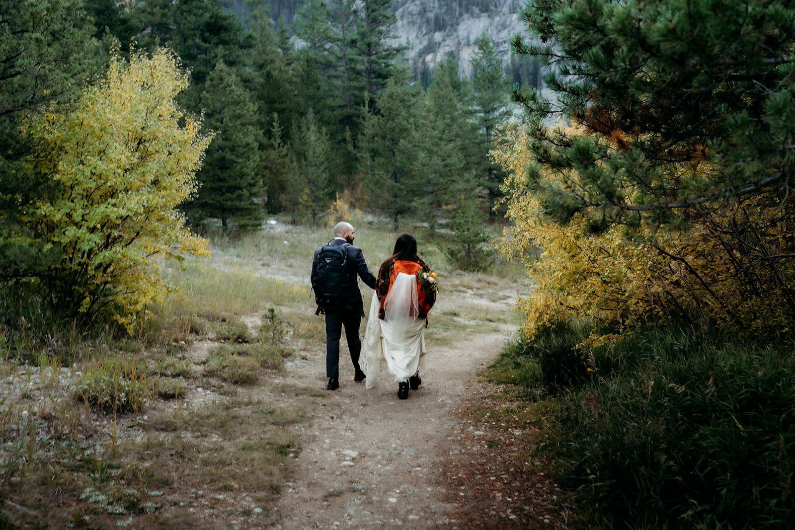 The newlyweds make their way down a quiet forest path, backpacks strapped over their wedding clothes, embodying the spirit of a true Colorado adventure elopement.