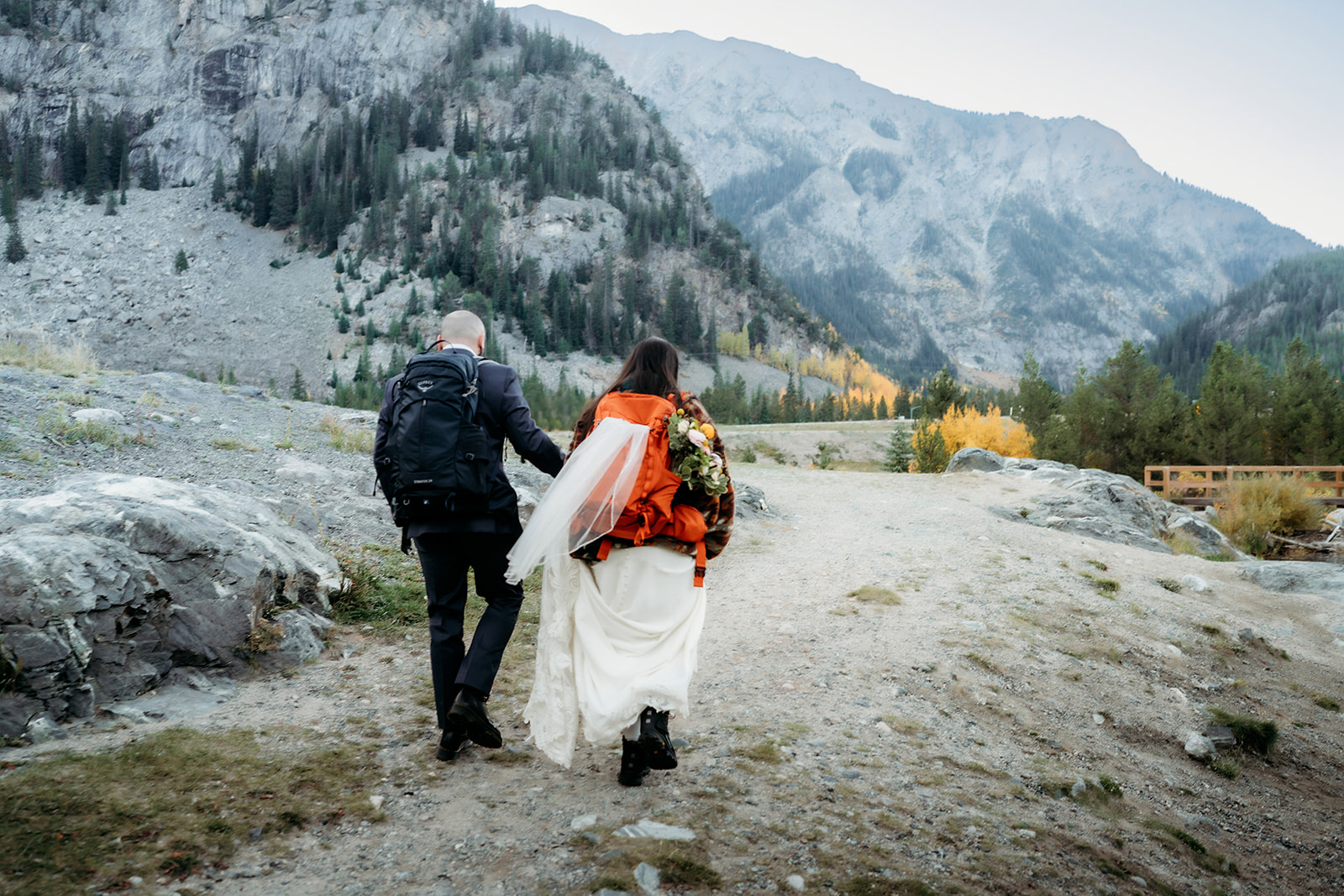 A bride and groom begin their adventure elopement by trekking along a rugged trail with backpacks and hiking boots beneath towering mountain cliffs.