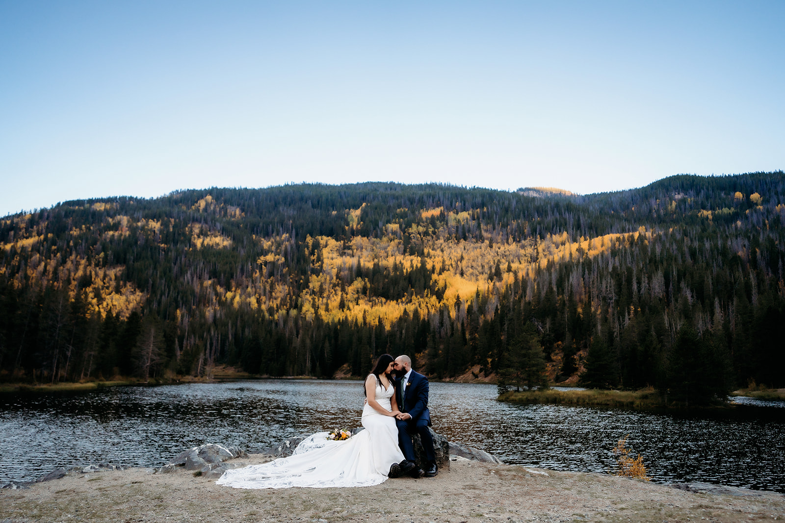 A newlywed couple shares an intimate moment beside an alpine lake surrounded by golden aspens and evergreens, framed by Colorado's fall foliage.