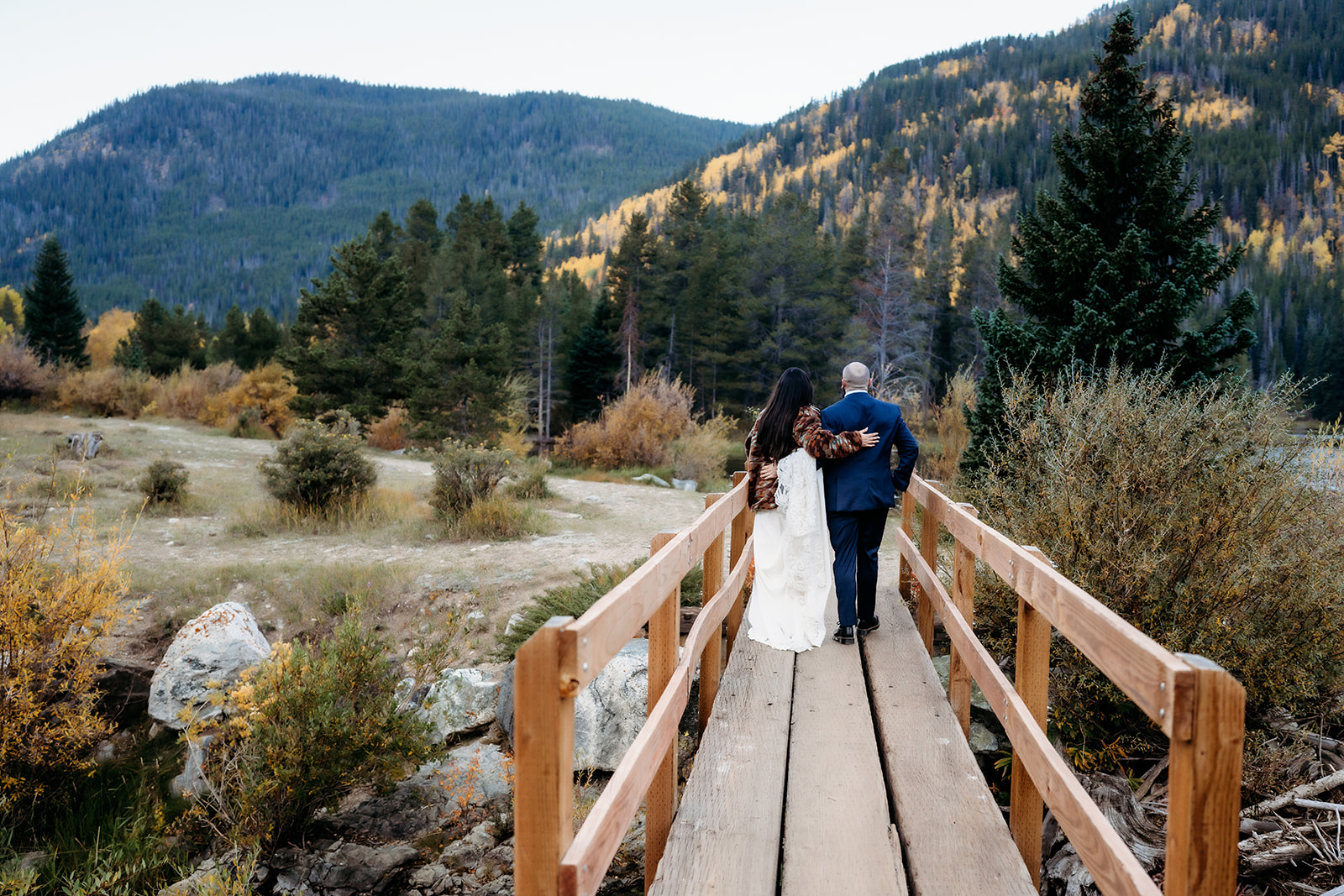 A bride and groom walk arm in arm across a wooden bridge, with a sweeping mountain view and warm autumn colors surrounding them—pure Colorado adventure elopement magic.