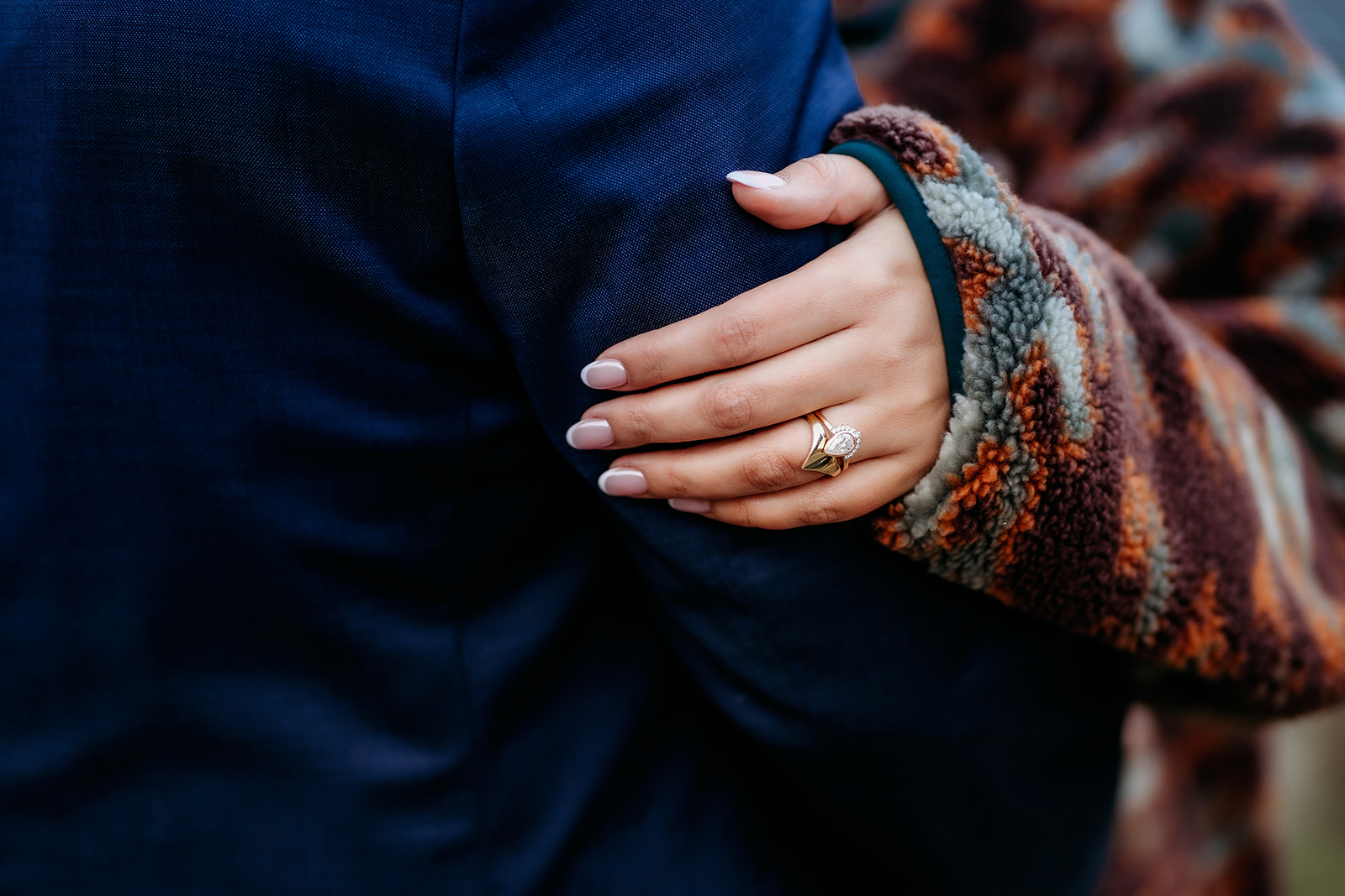 A detailed shot of the bride’s engagement and wedding rings as she gently holds her groom’s arm, her hand wrapped in a warm patterned sleeve.