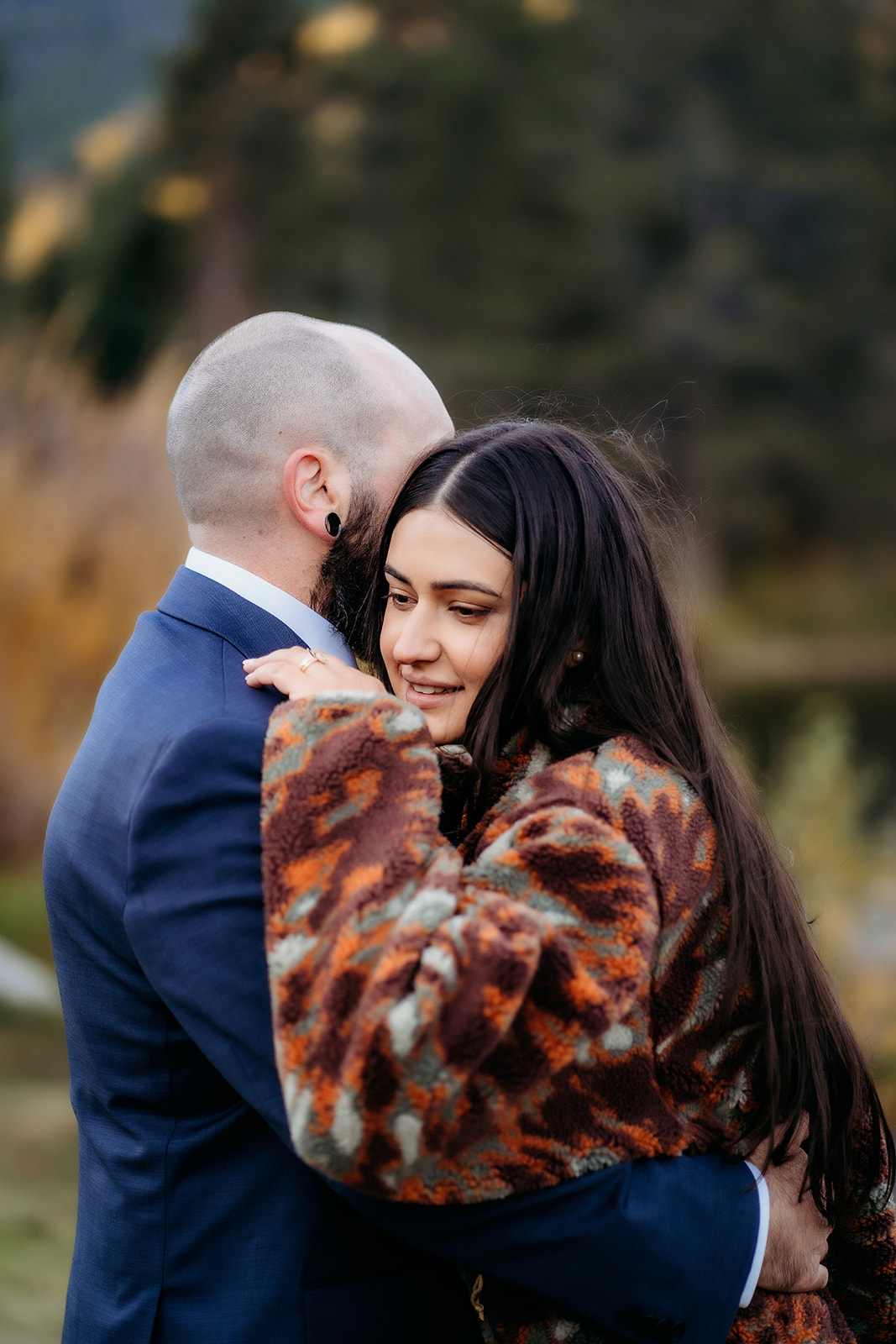 A bride wrapped in a warm patterned coat smiles softly while hugging her groom during a quiet mountain moment in their elopement celebration.
