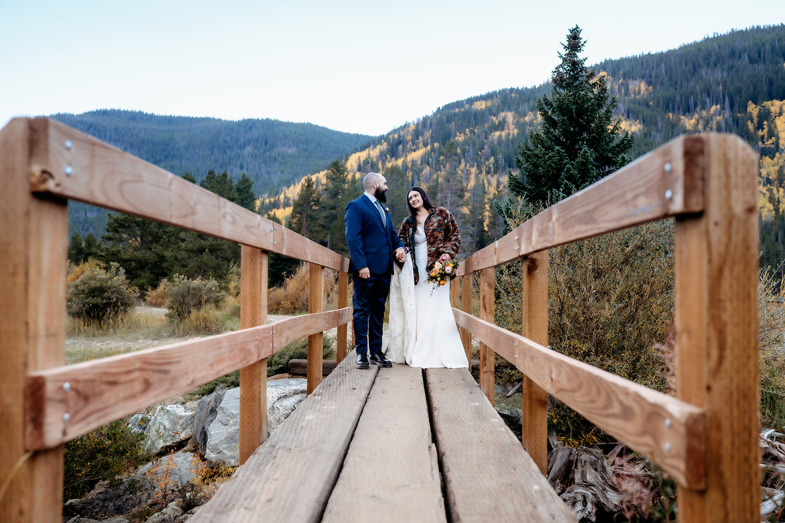 The bride and groom pause mid-bridge, looking at one another with big smiles as mountains and autumn foliage surround them—capturing the heart of an adventure elopement in Colorado.