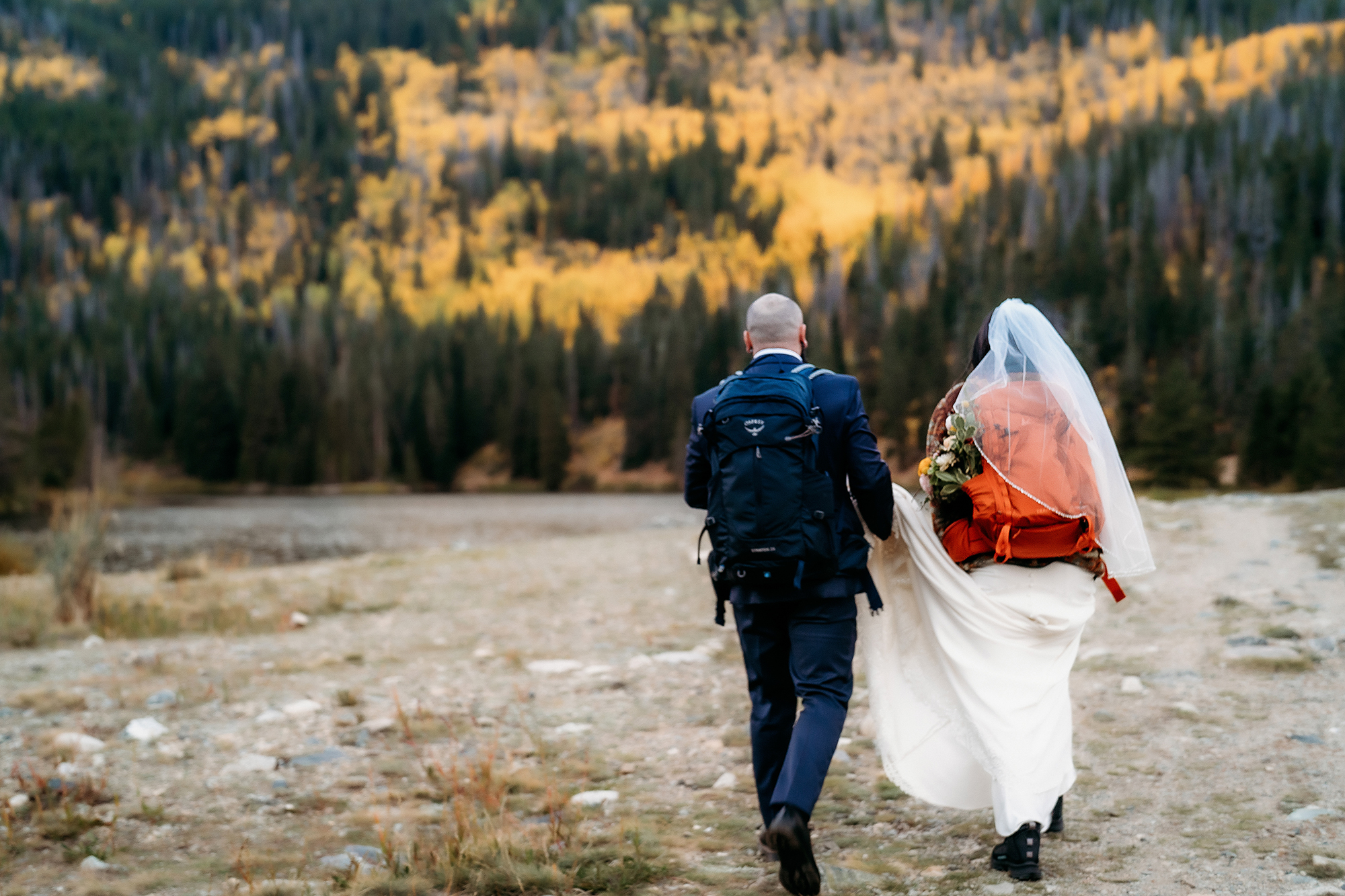 The couple hikes toward the glowing aspen-covered mountains, backpacks full, veil blowing in the wind—capturing the essence of an authentic Colorado adventure elopement.