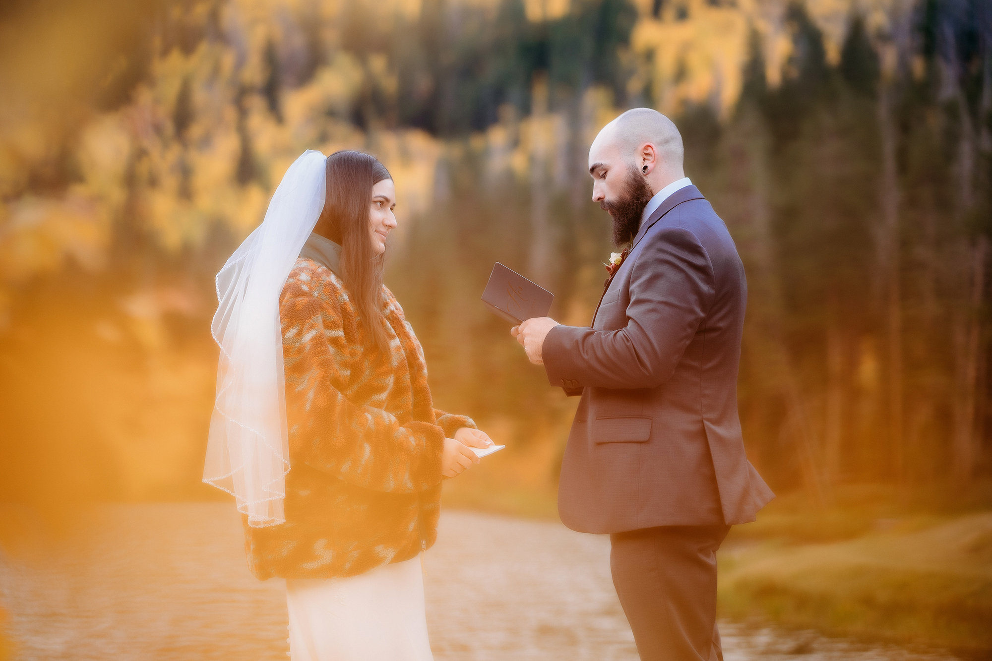 Soft golden leaves frame a heartfelt moment as the groom reads vows to his bride during their private adventure elopement ceremony by the lake.