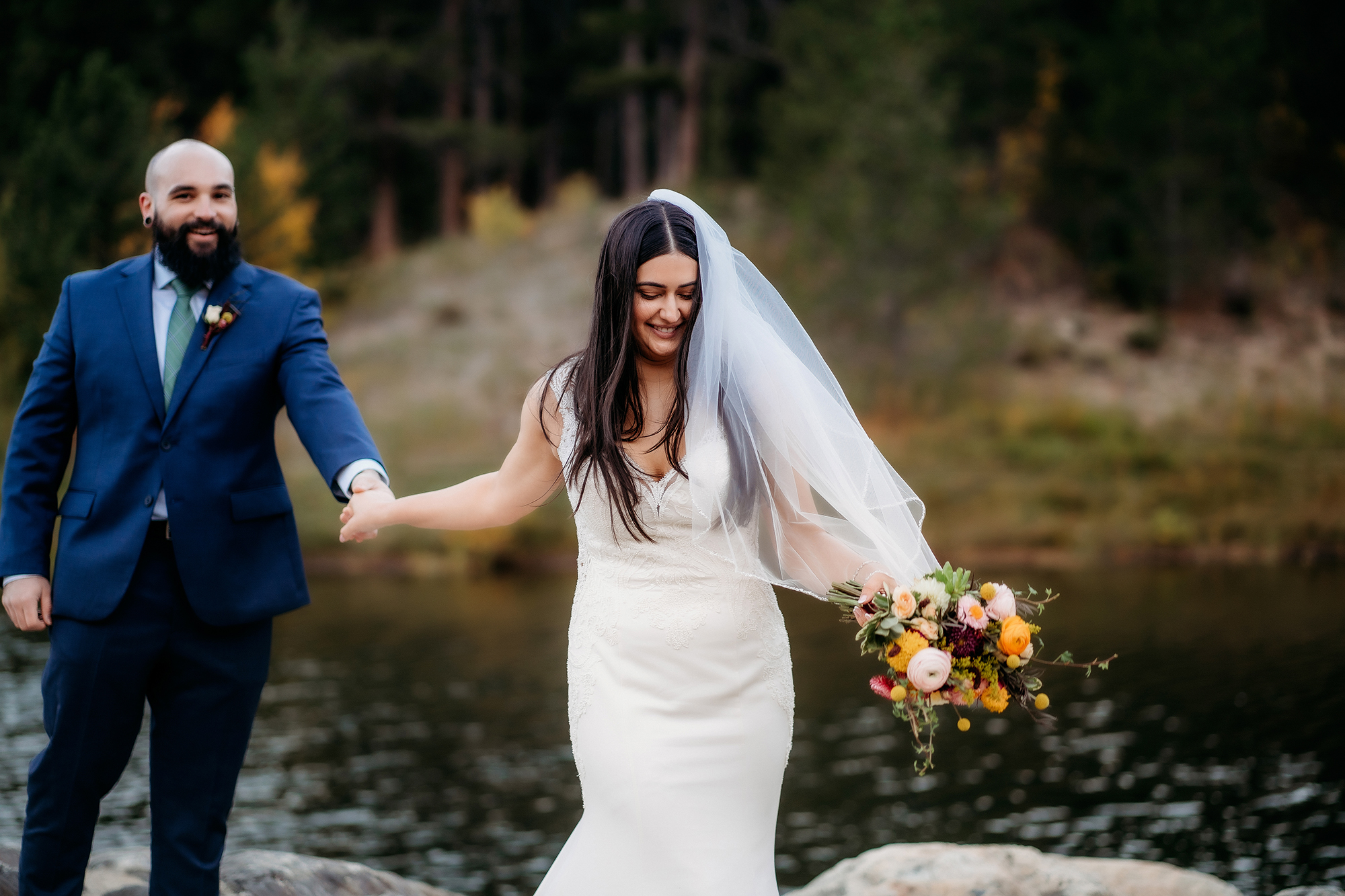 A joyful bride in her wedding gown and veil leads her smiling groom along the lakeshore, bouquet in hand and laughter in the air.