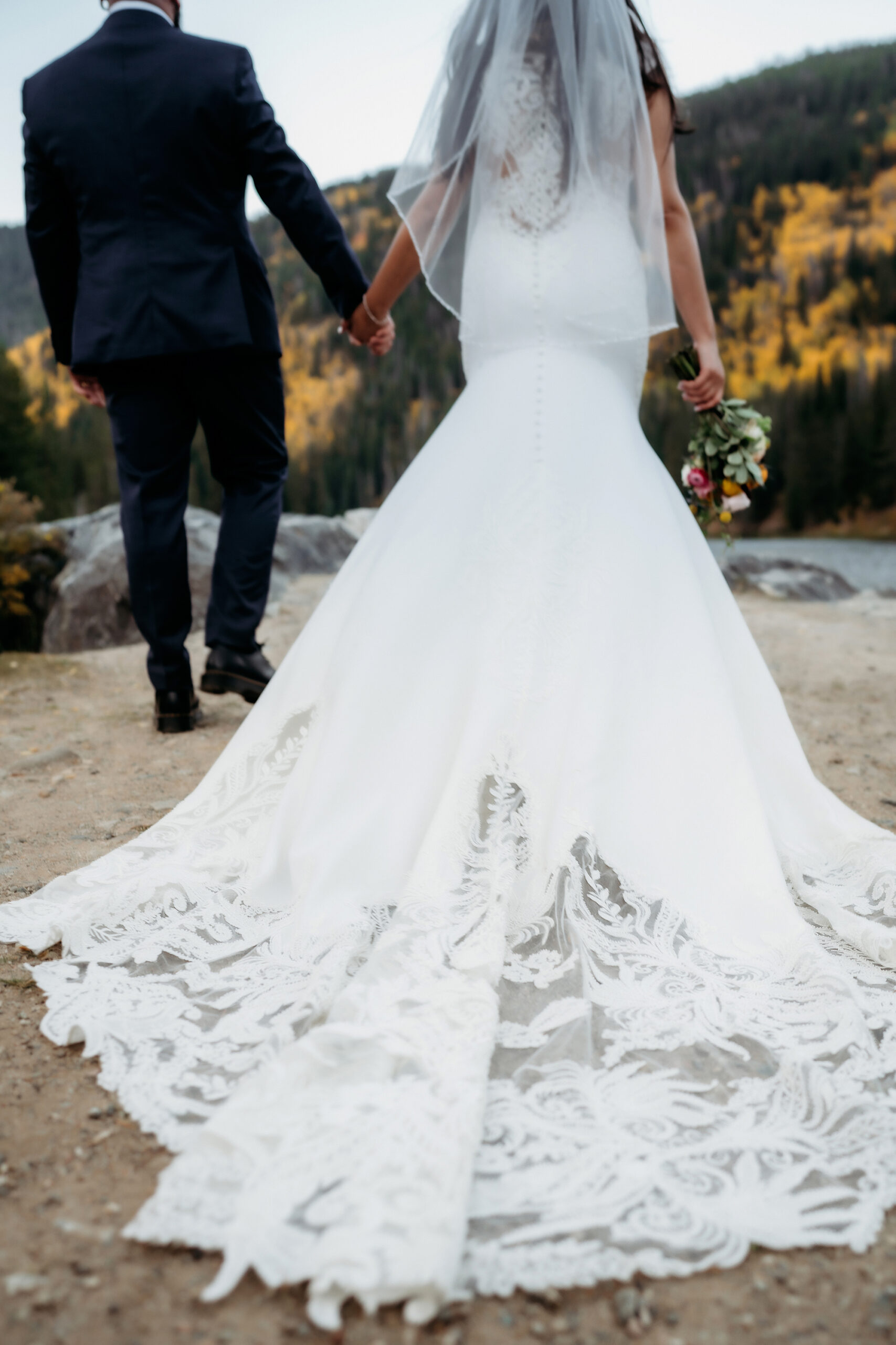The intricate lace train of the bride’s gown trails behind her as she walks hand in hand with her partner toward a scenic mountain overlook.