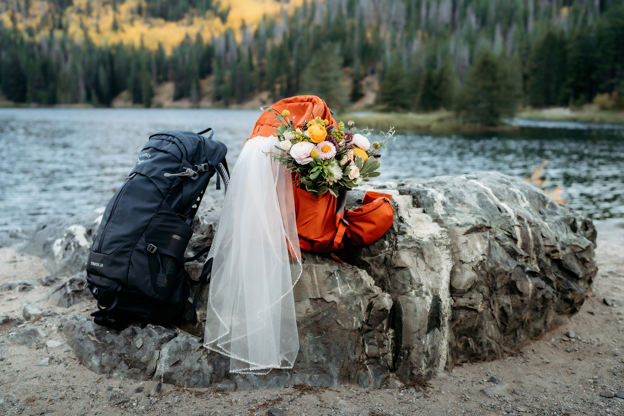 Two hiking backpacks rest on a boulder—one draped with a wedding veil and bouquet—symbolizing the beauty and spirit of an adventure elopement in the Colorado wilderness.