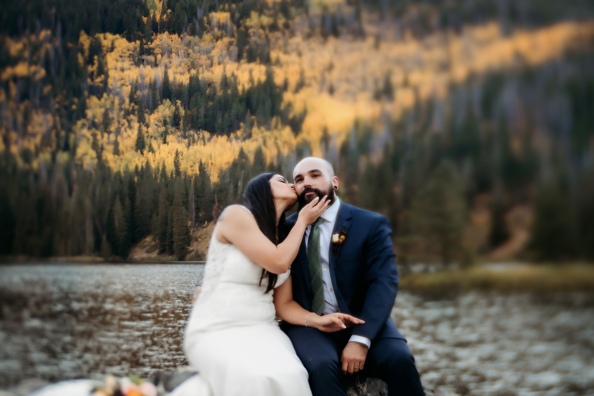 A playful moment unfolds as the bride leans in to kiss her groom’s cheek while they sit together near a lake glowing with fall color.