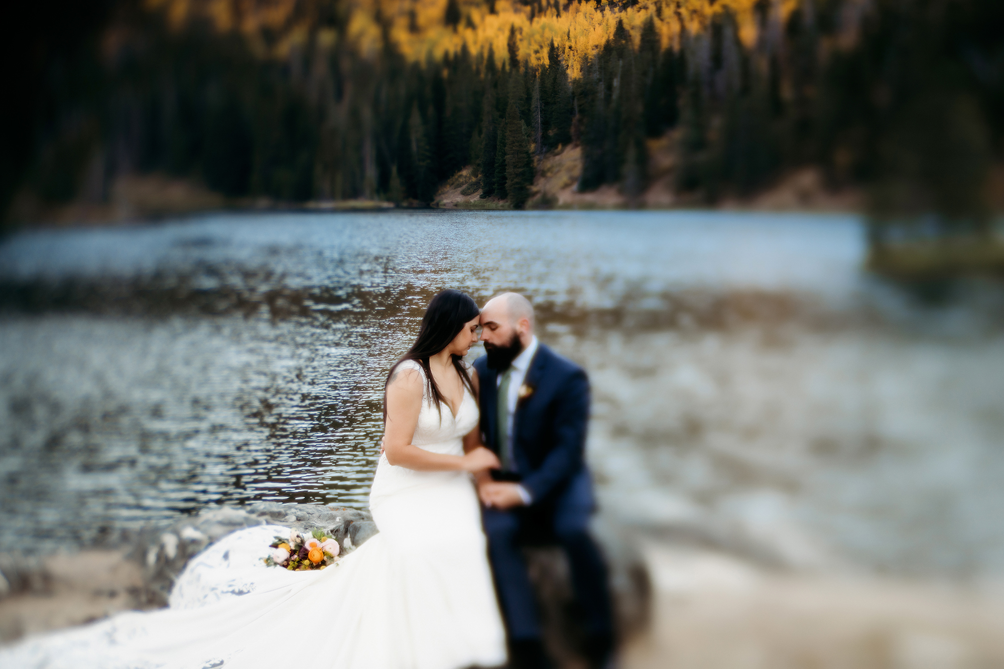 A bride and groom share a quiet, heartfelt moment by the water’s edge, their foreheads gently touching, framed by the glowing foliage of their fall adventure elopement.