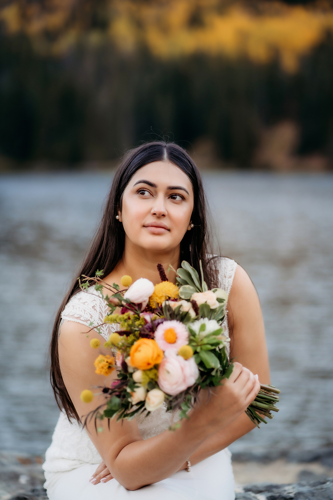 A bride sits beside a lake holding a vibrant bouquet of wildflowers, her gaze dreamy and serene against a backdrop of autumn colors.