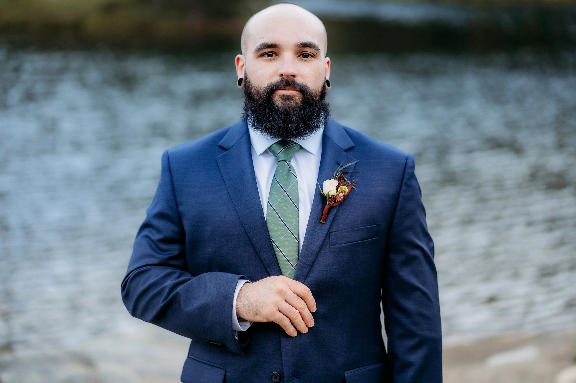 A groom stands confidently in a navy suit and plaid green tie, boutonnière pinned neatly, with the still waters of a Colorado lake behind him.