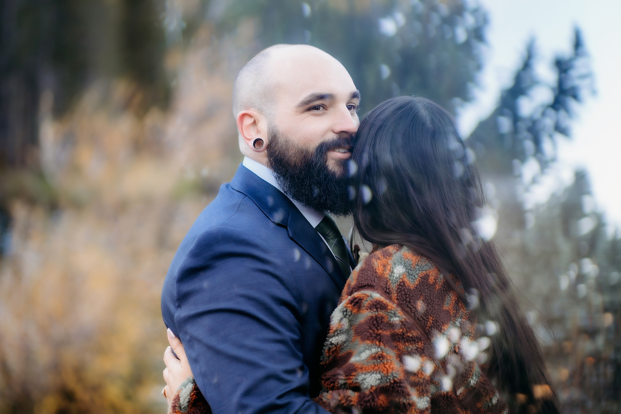 The groom smiles softly as he’s embraced by his bride in a cozy patterned jacket, surrounded by dreamy fall foliage and soft reflections during their adventure elopement.