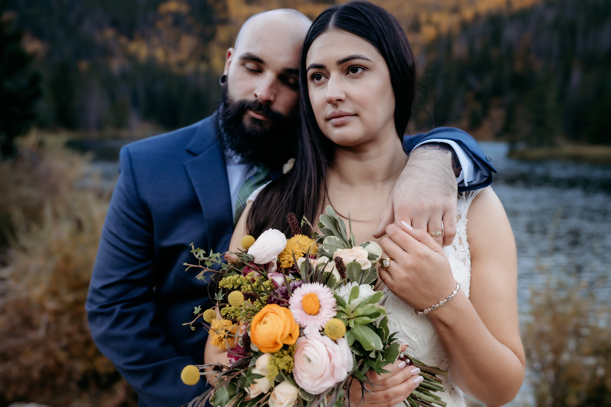 A tender moment as the groom closes his eyes and rests his head on the bride’s shoulder, her bouquet bursting with fall florals beside a tranquil lake.