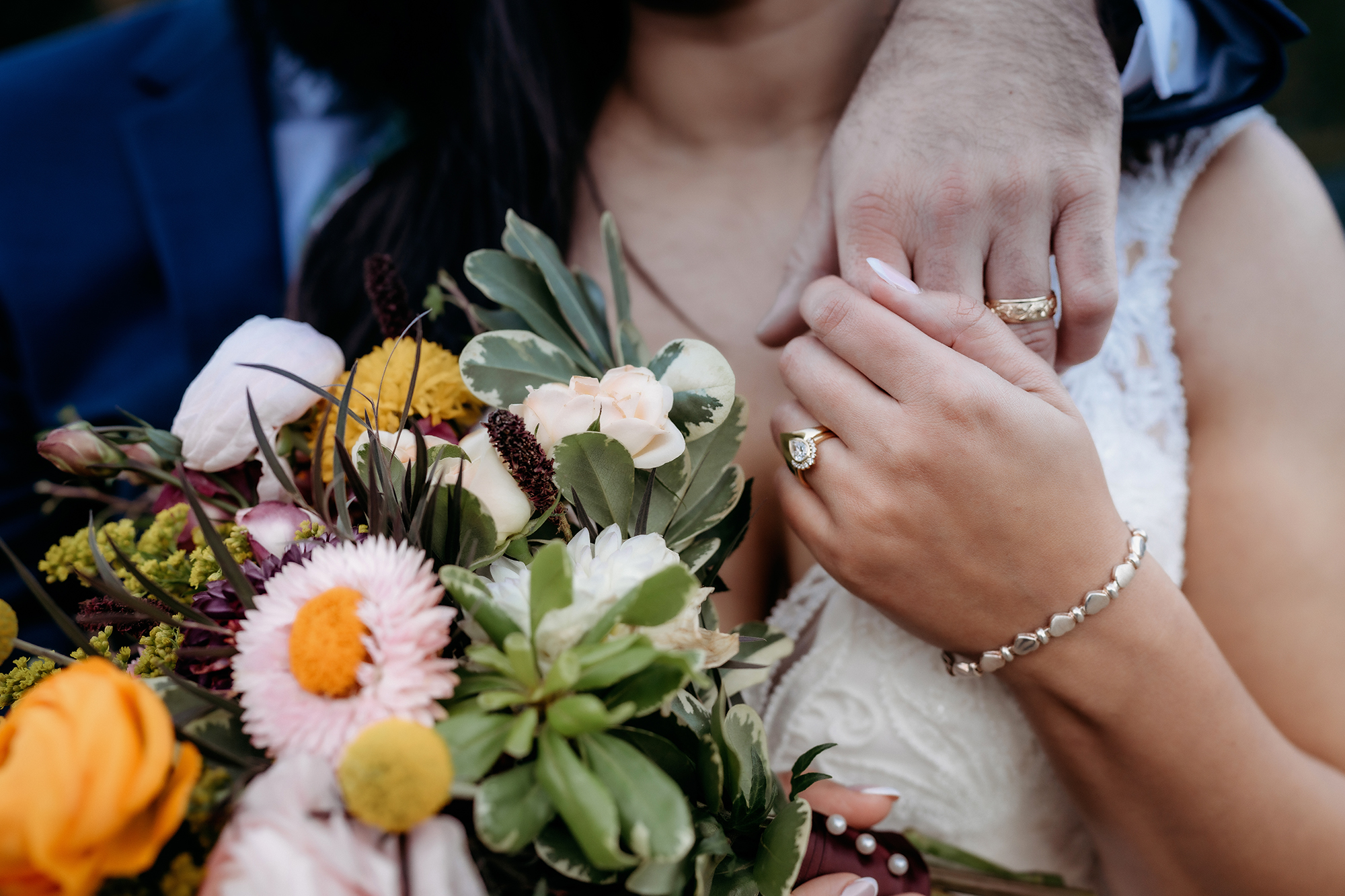 A close-up of the couple’s intertwined hands, showing matching rings and a bright, wildflower-inspired bouquet bursting with texture and color.