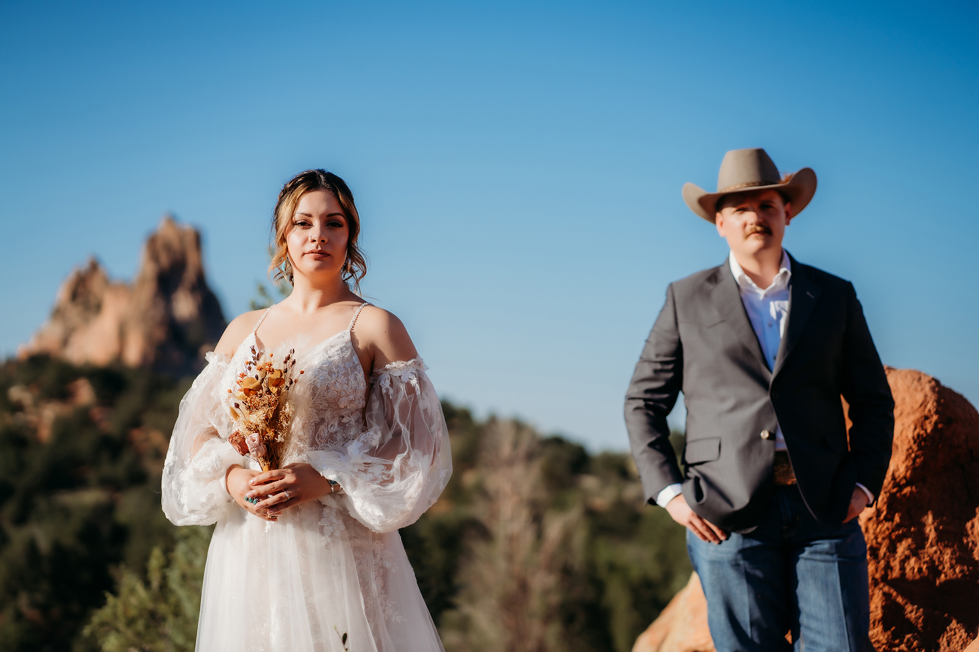 Bride holding a rustic bouquet standing confidently in a white lace wedding dress with groom wearing a cowboy hat in the background at a Denver elopement.