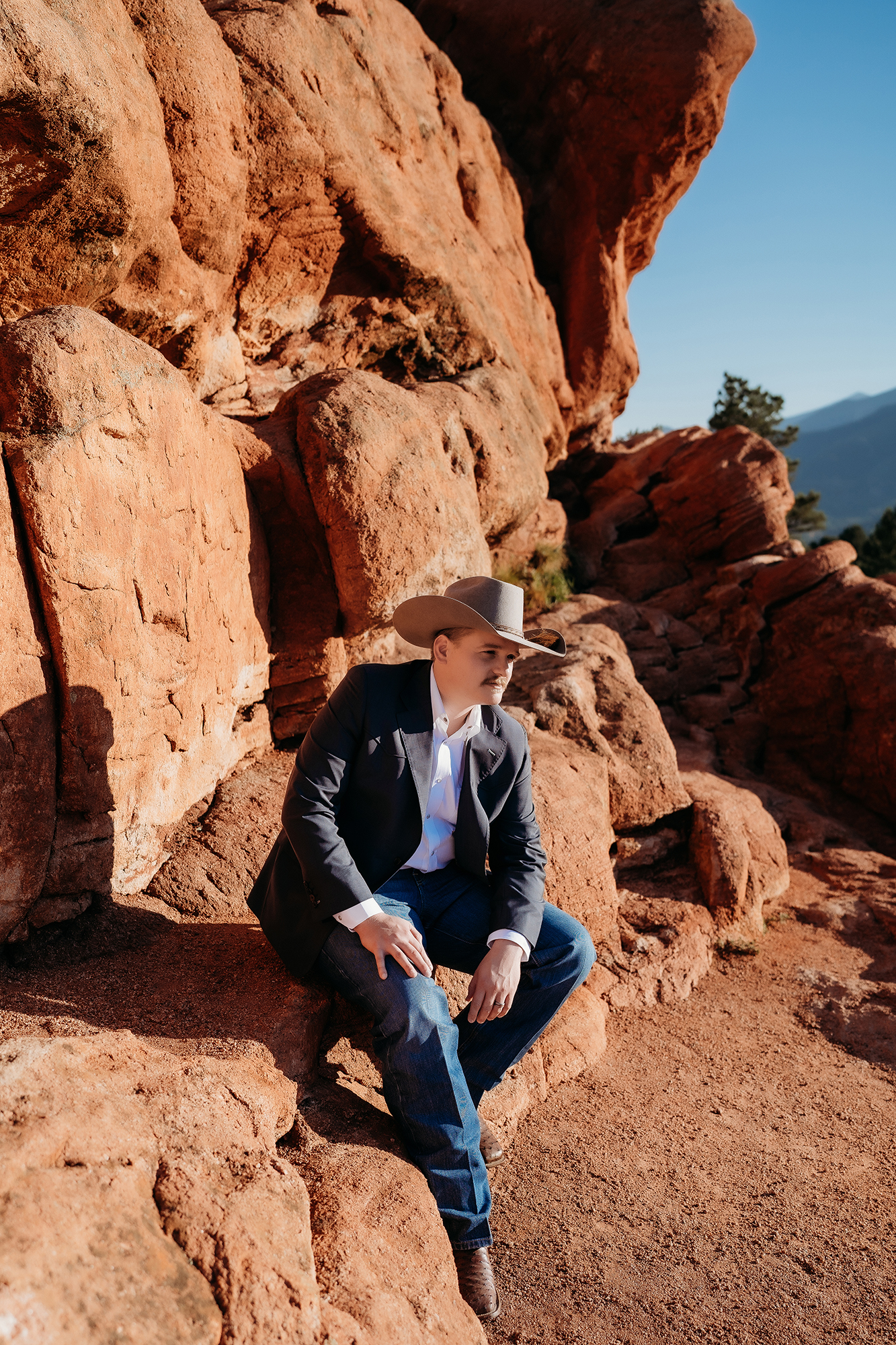 Groom sitting thoughtfully on red rocks wearing cowboy hat during a Denver elopement photoshoot.