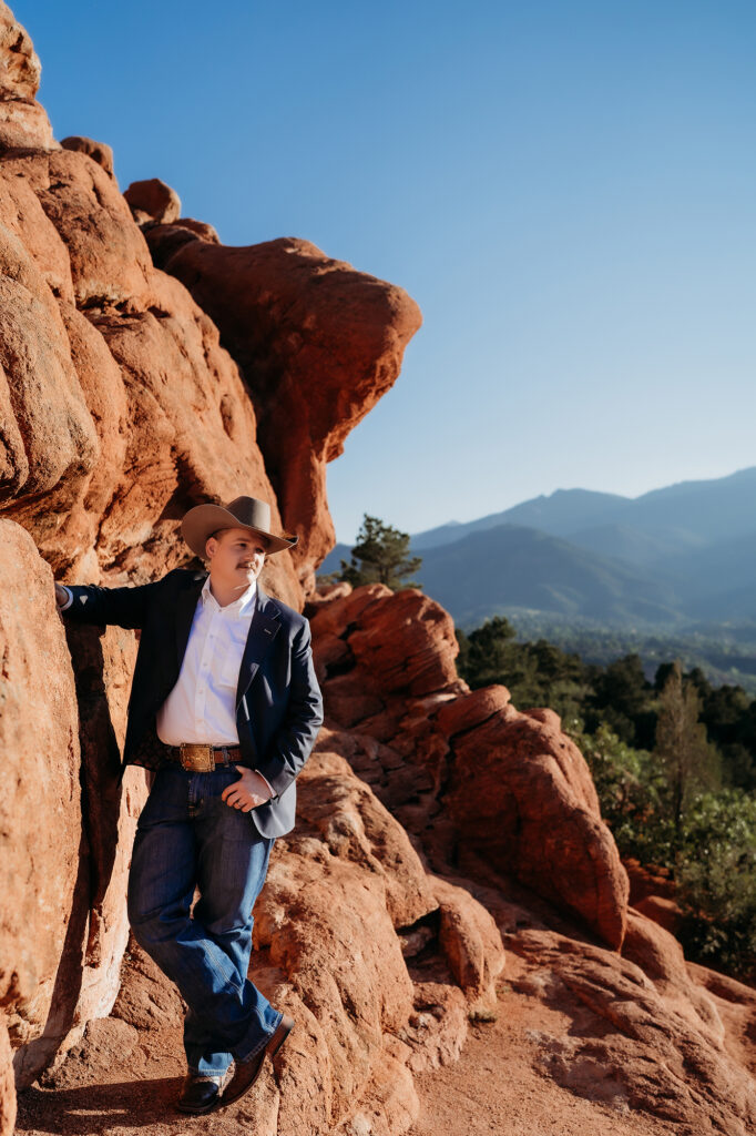Groom wearing cowboy hat and blazer leaning against red rock formations with mountain views during a Denver elopement session.