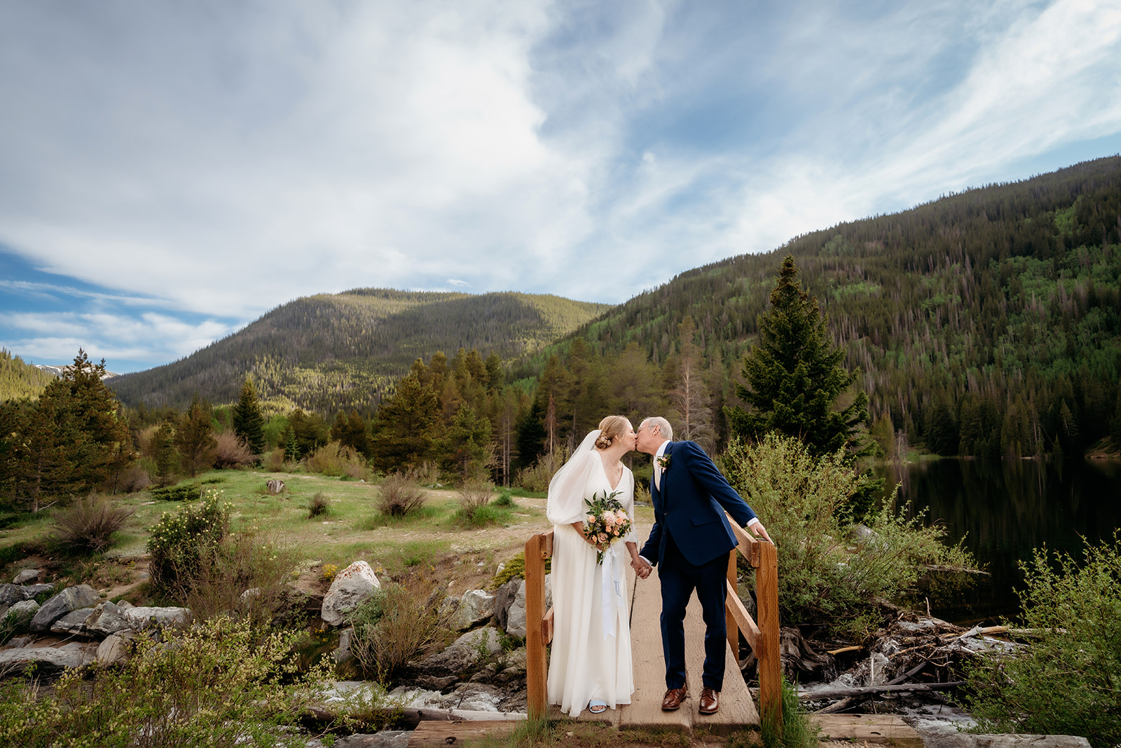 The couple shares a romantic kiss on a rustic bridge, with forested hills behind them—capturing the joy of a private elopement.