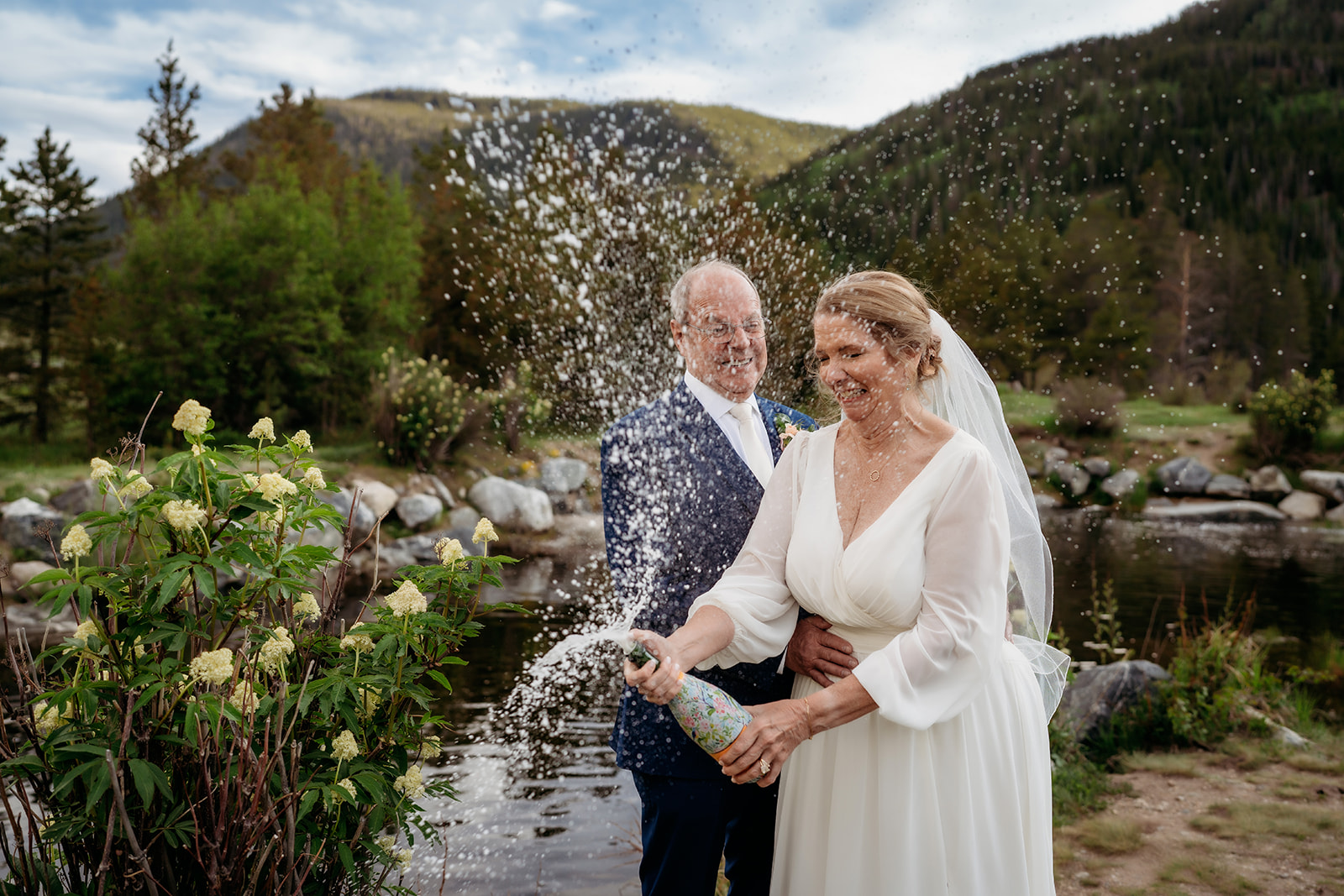 The bride joyfully sprays champagne as her groom stands behind her, both laughing beside a mountain pond—an unforgettable way to celebrate How to Make Your Elopement Special.