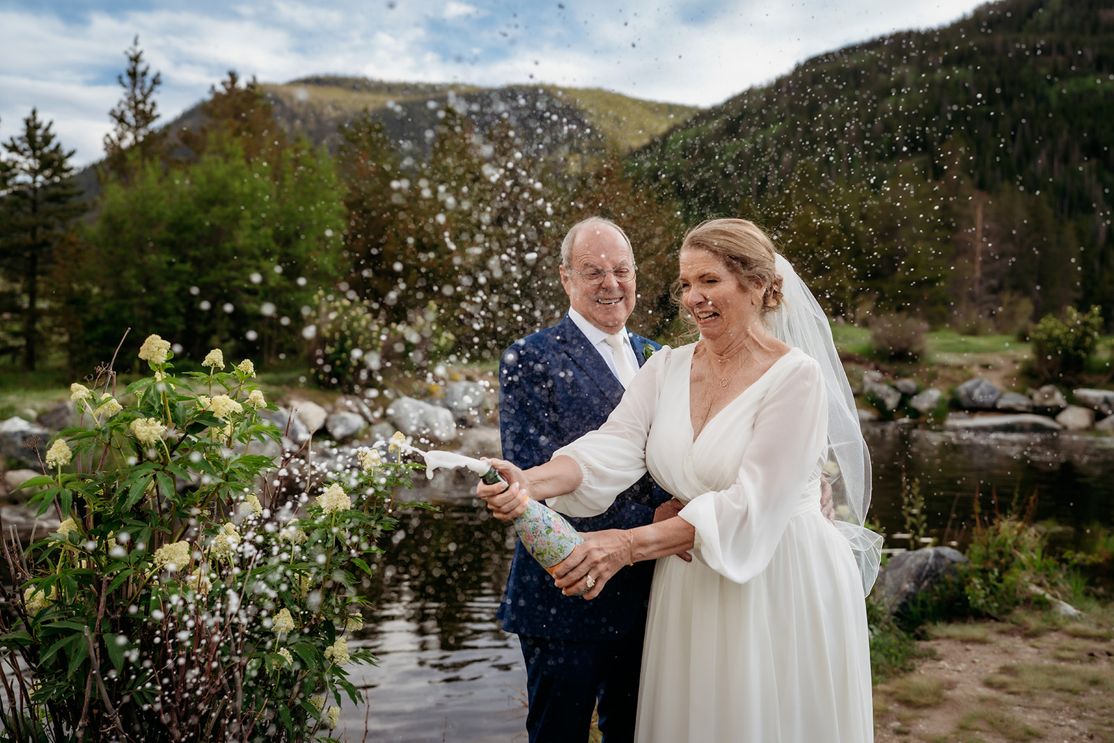 Laughing and carefree, the couple pops a champagne bottle beside a scenic mountain pond.