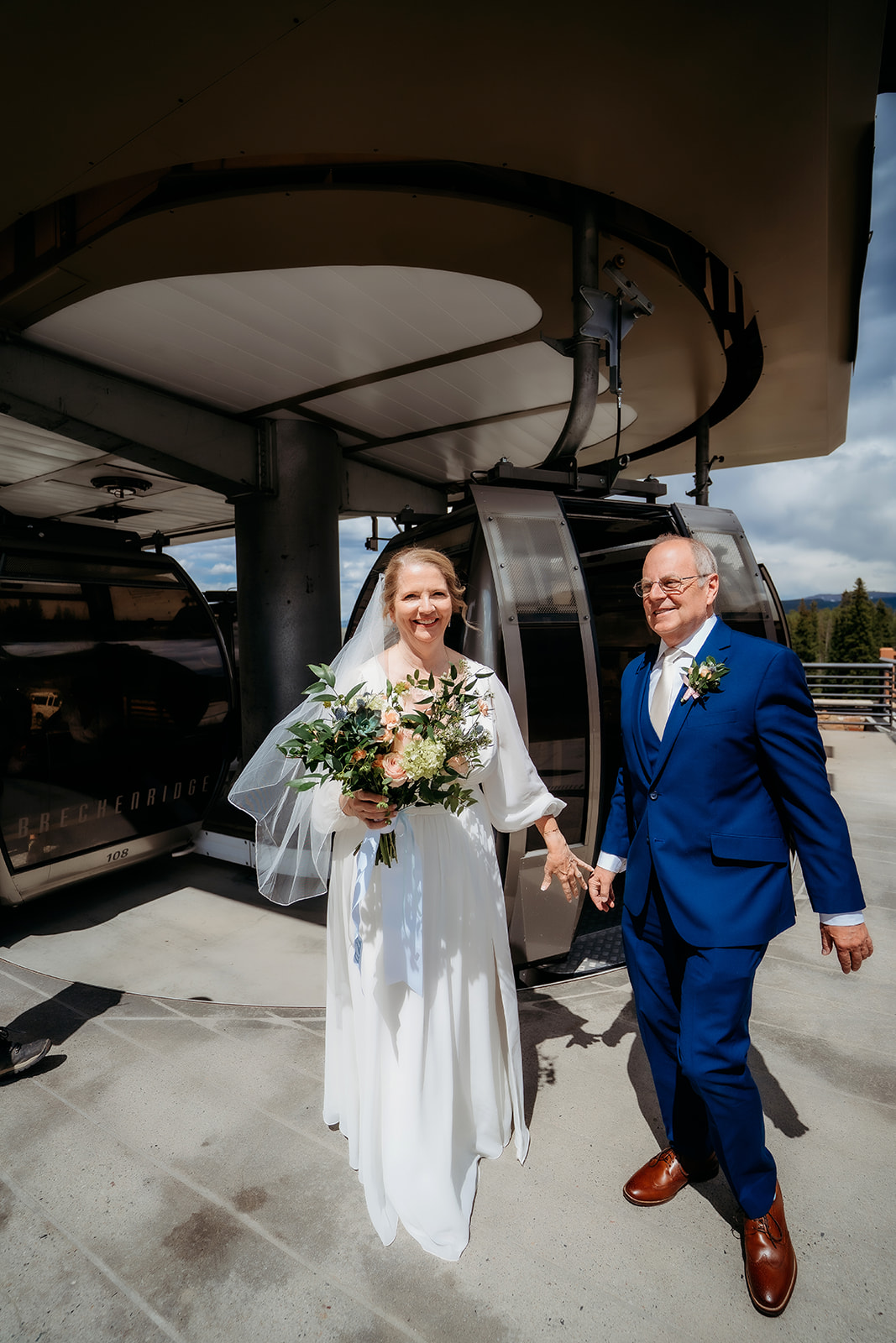 Smiling and hand-in-hand, the bride and groom arrive at a gondola station for a scenic ascent on their wedding day.