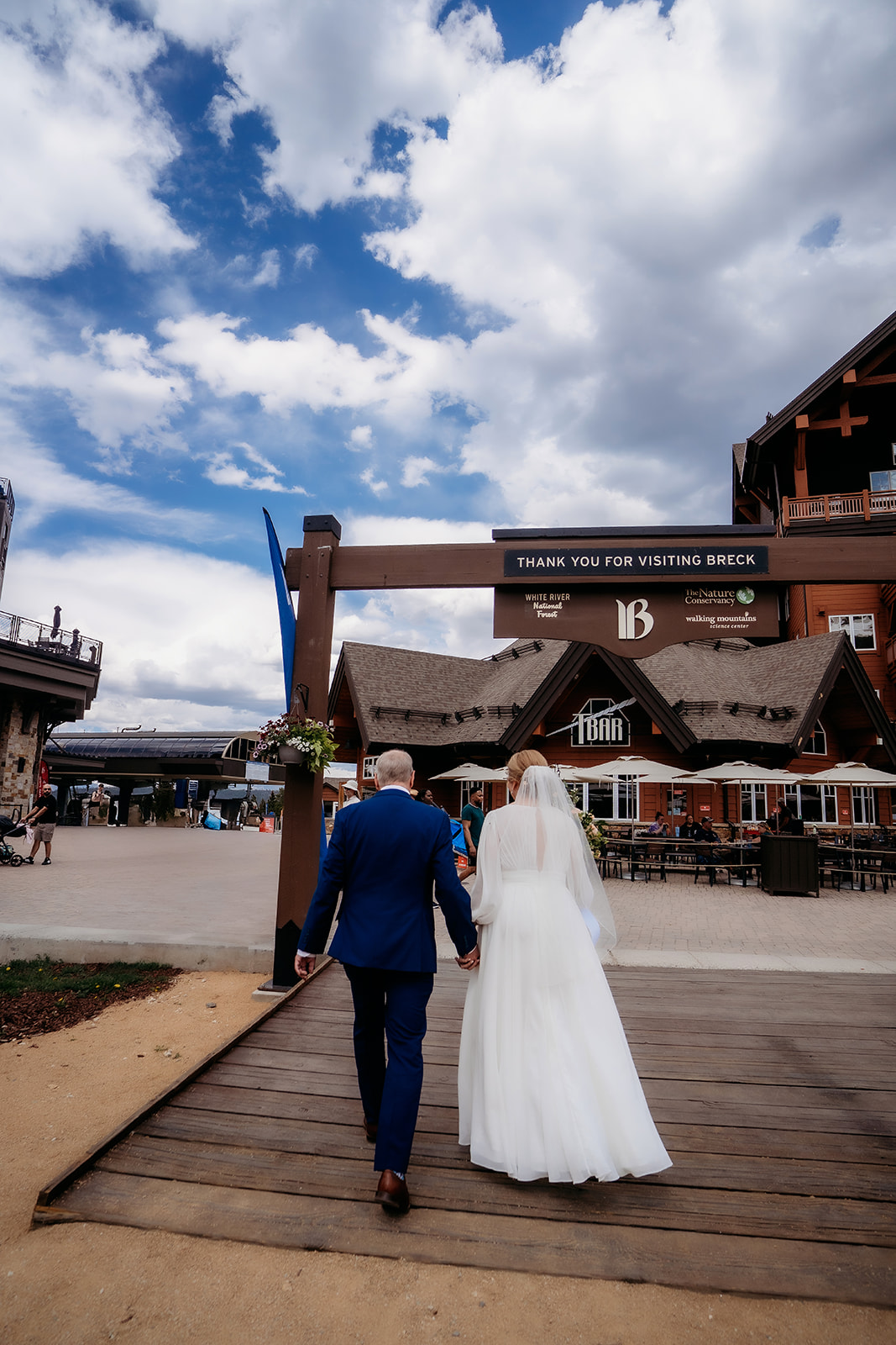 The newlyweds stroll beneath a wooden “Thank You For Visiting Breck” sign—marking the end of a joyful, scenic celebration.