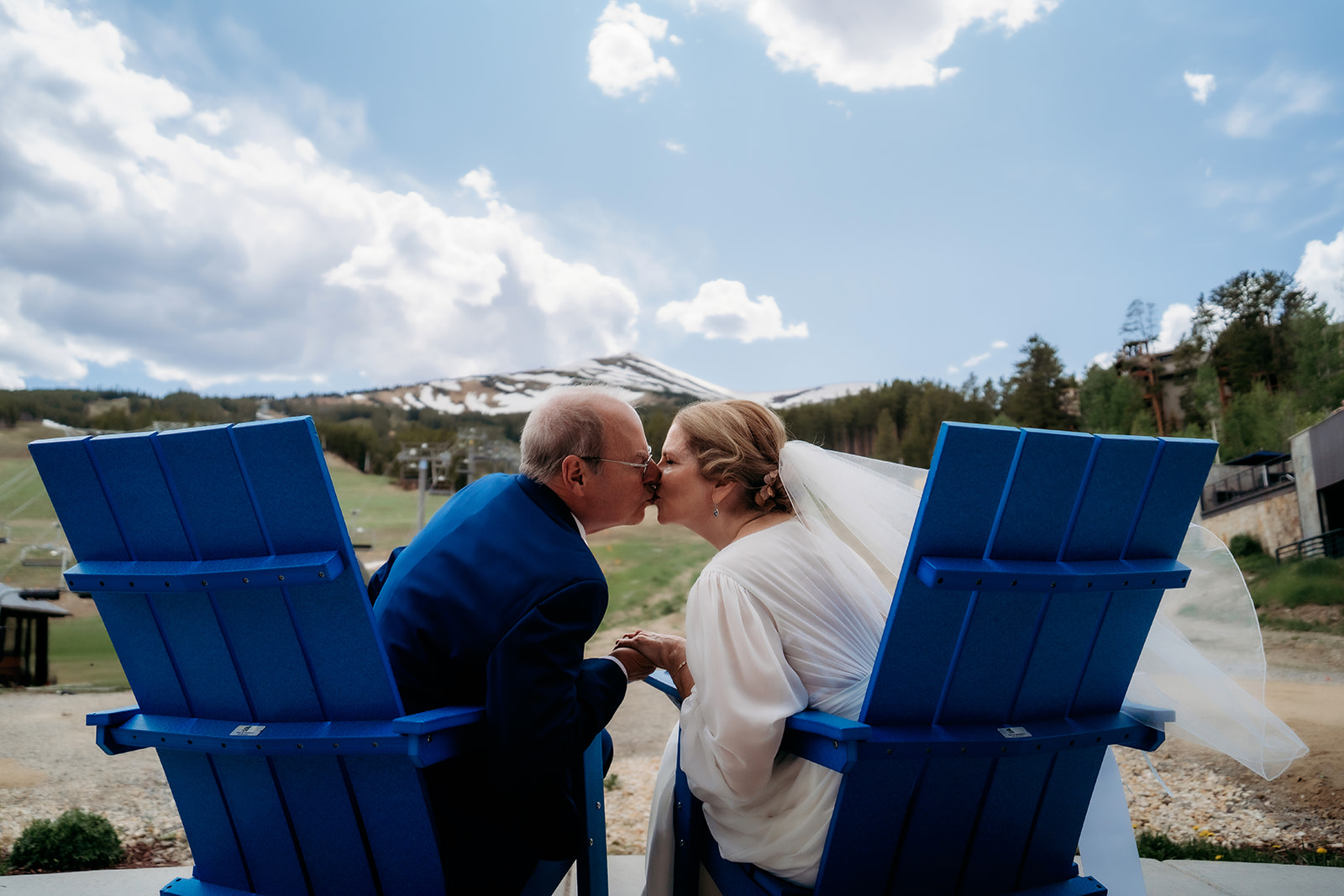 Seated in matching blue chairs, the couple shares a kiss with mountain slopes behind them—an intimate pause that reflects How to Make Your Elopement Special.